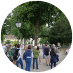 Eine Gruppe von Menschen steht mit dem Rücken zur Kamera unter ein paar Bäumen und hört einem Mann zu.