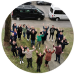 Eine Gruppe von Menschen steht in Herzform auf einer Rasenfläche und hebt die Hände nach oben in die Kamera.
