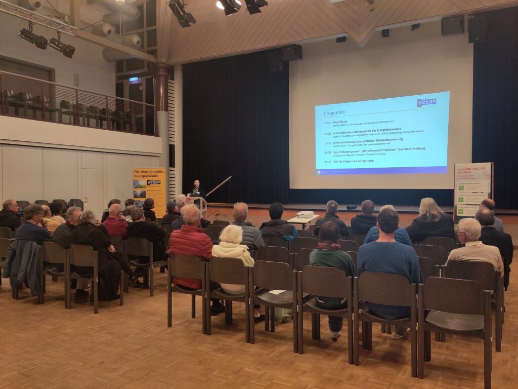 Publikum sitzt in einem Saal und sieht eine Präsentation mit Programm auf einer Leinwand.