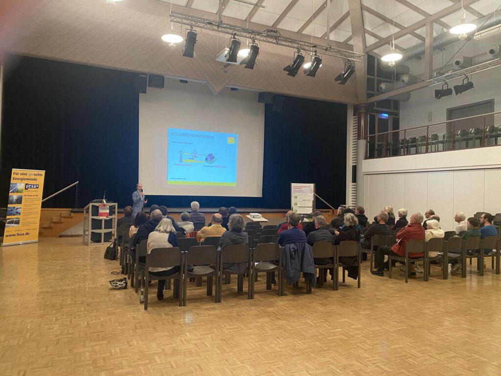 Vortragender steht vor Leinwand mit Präsentation, Publikum sitzt auf Stühlen in großer Halle mit Holzfußboden und hoher Decke.