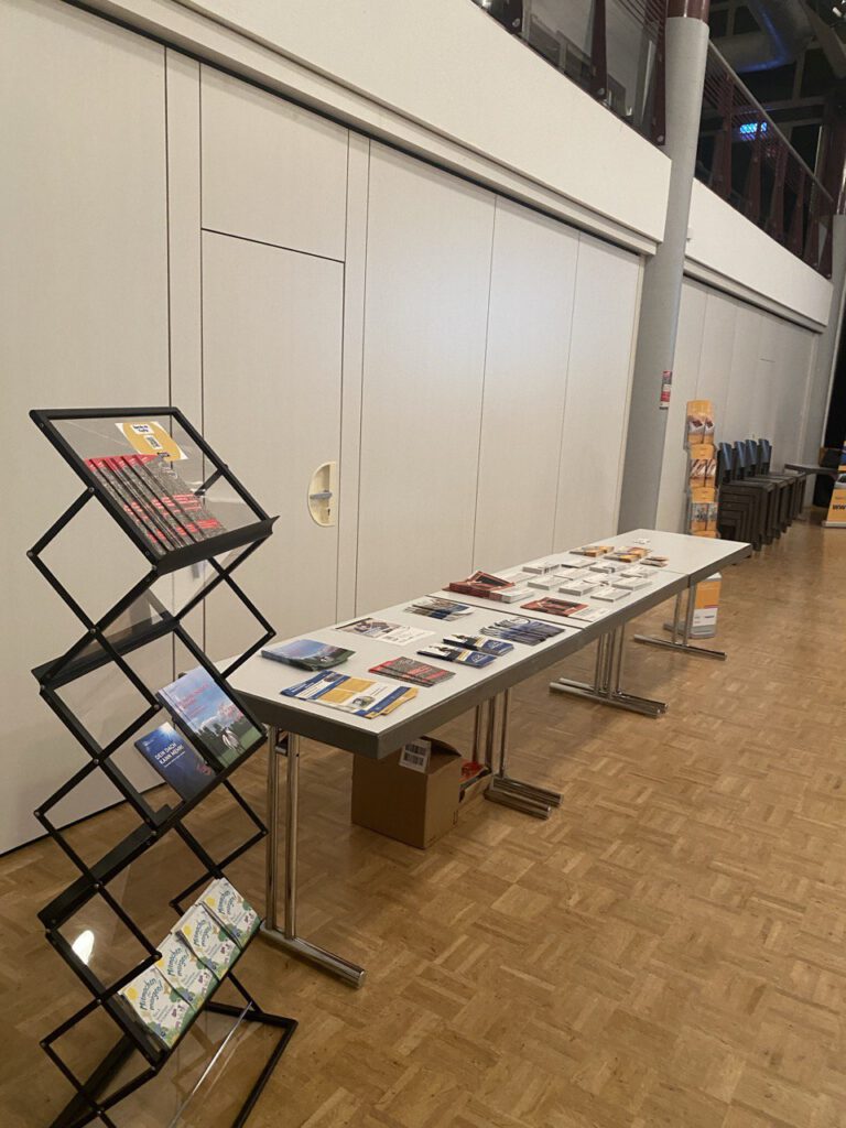Langer Tisch mit verschiedenen Flyern und Broschüren, daneben ein schwarzes Prospektständer mit weiteren Informationsmaterialien in einem großen Raum mit Holzfußboden