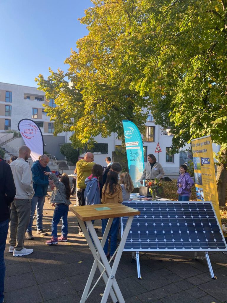Klimaquartier Auftakt: Menschen stehen an dem Infostand von fesa mit Solarpanelen und bunten Fahnen vor einem Baum und Wohngebäuden