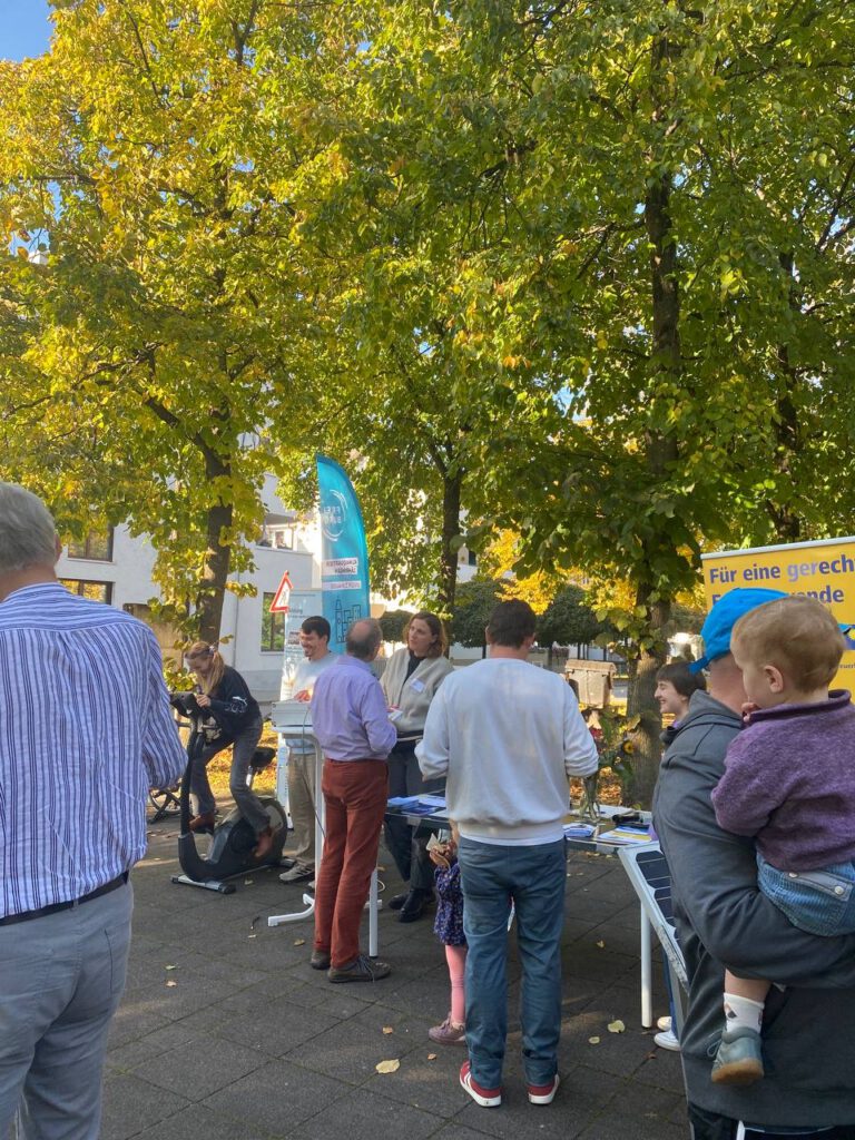 Klimaquartier Auftakt: Menschen stehen an einem Infostand im Freien unter Bäumen mit grünen Blättern, ein Kind sitzt auf dem Arm eines Erwachsenen.
