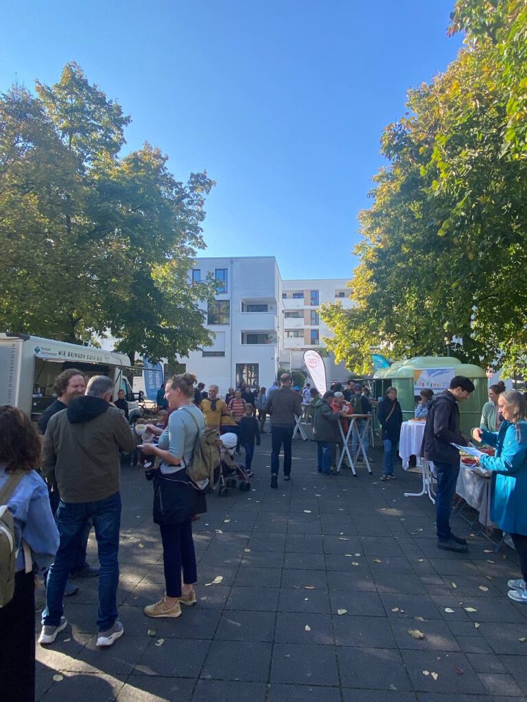 Klimaquartier Auftakt: Menschen stehen an Ständen und Tischen im Freien auf einem gepflasterten Platz zwischen Bäumen und einem weißen Wohngebäude im Hintergrund.