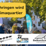 Menschen bei einer Veranstaltung im Freien mit Bannern und Schildern, darunter die Aufschriften 'Zähringen wird Klimaquartier', 'energiekarawane', 'Mein Strom vom Balkon' und 'BalkonScout Freiburg'.