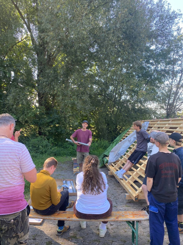 Die Teilnehmenden stehen neben einem Dachgestell aus Holz. Der Workshop-Leiter erklärt ihnen etwas.